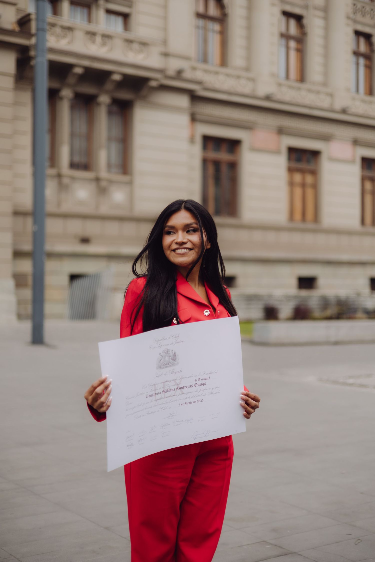 Smiling woman in red suit holding an official certificate outdoors in front of a historic building.