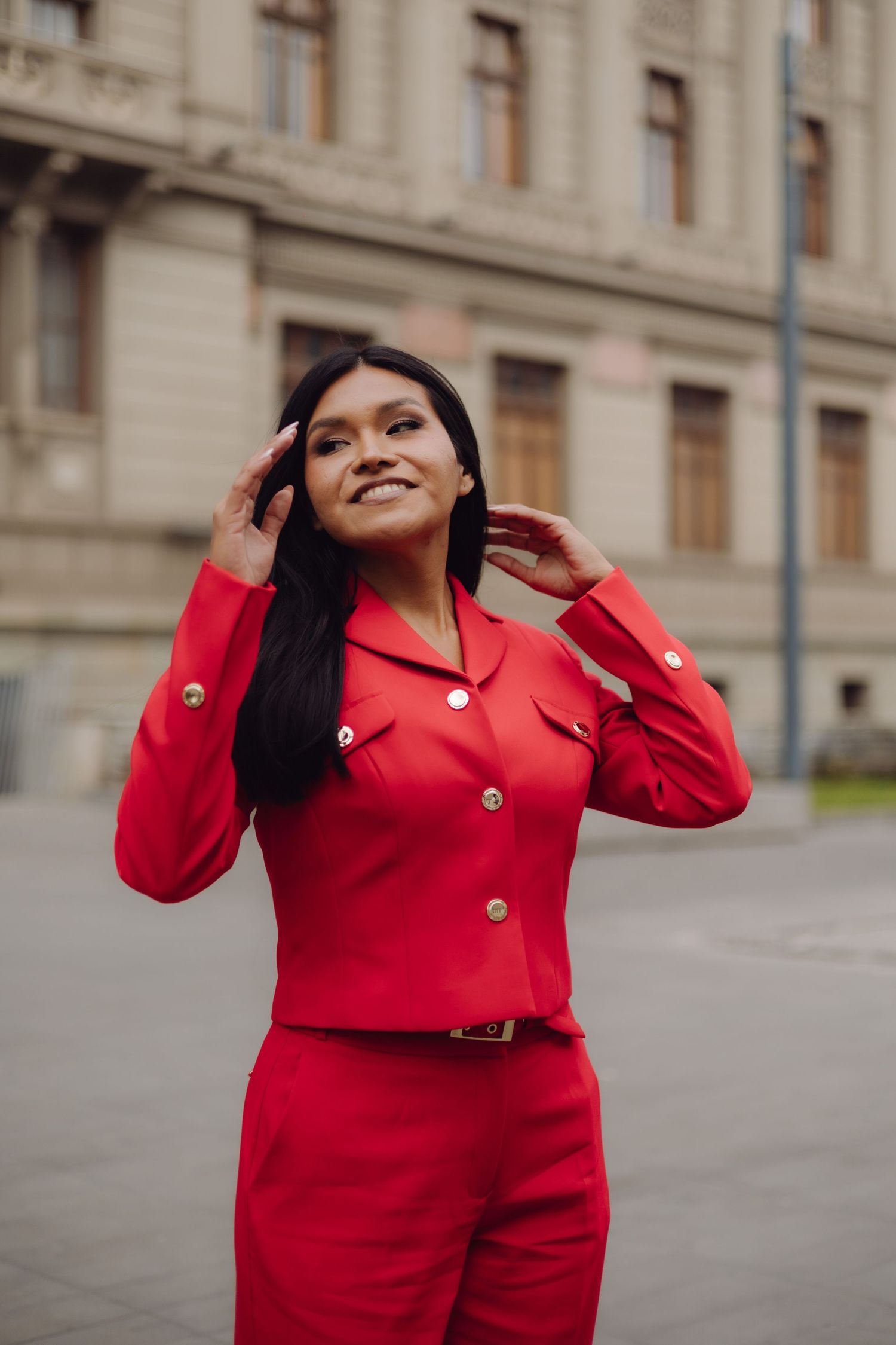 Smiling young woman in red suit adjusting hair outdoors near historic building.