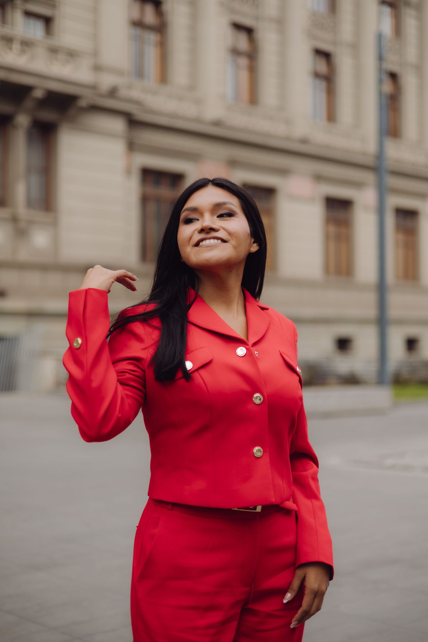 Happy young woman in red blazer and pants posing outdoors near classic building.
