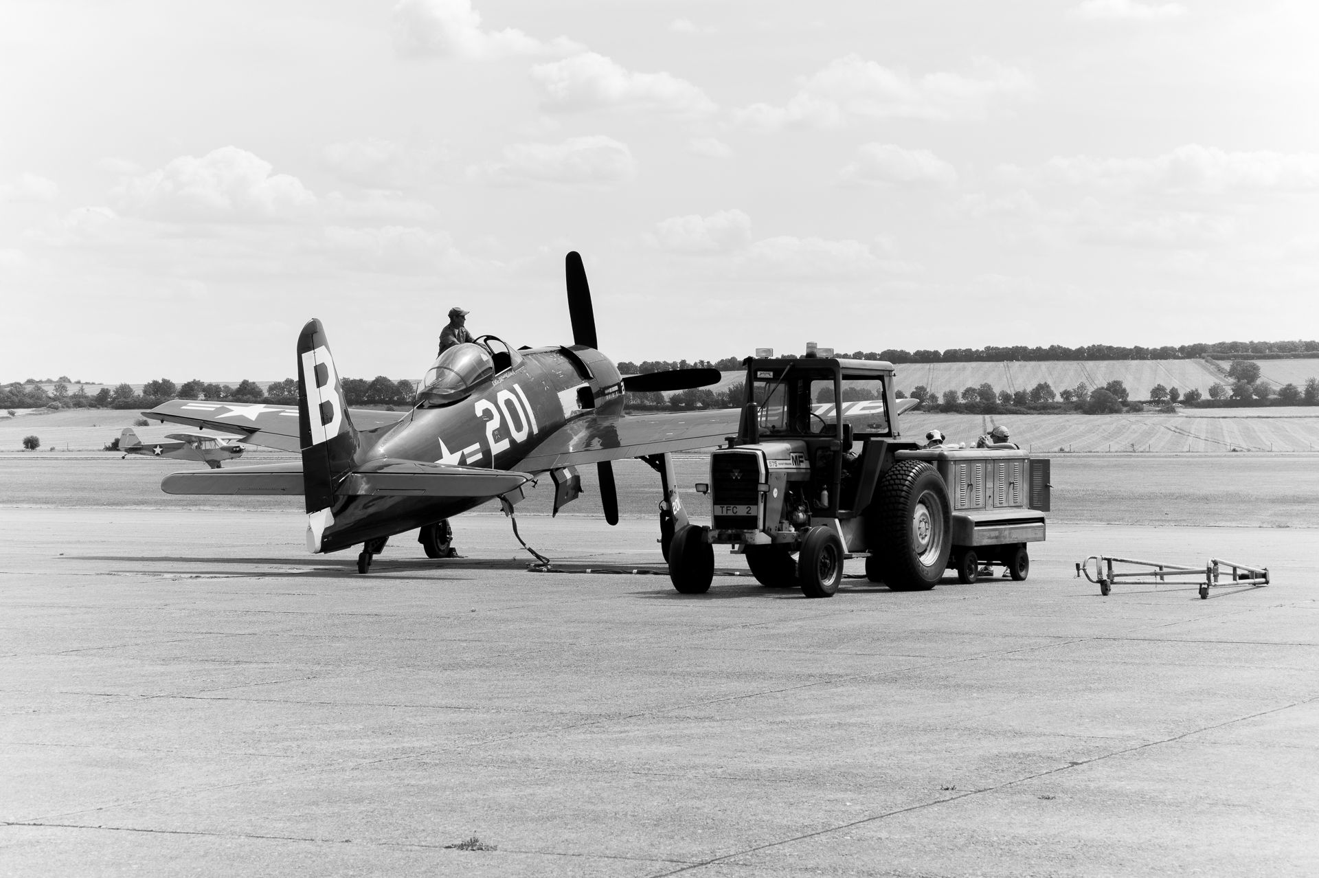 Vintage WWII fighter plane being towed by a tractor on an airfield runway in a black and white photograph.