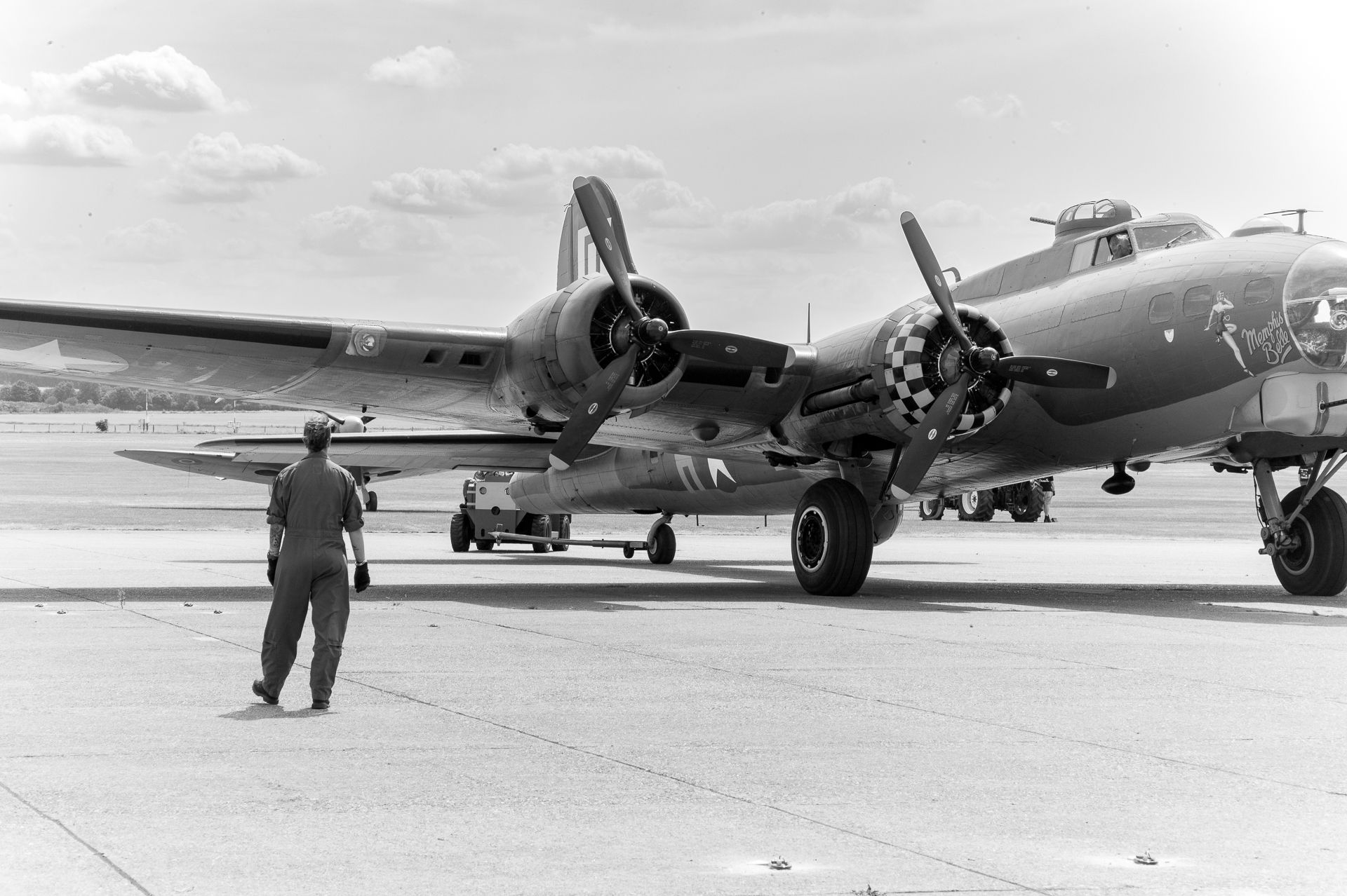 WWII-era B-17 Flying Fortress bomber on tarmac with ground crew member walking nearby, black and white photo.