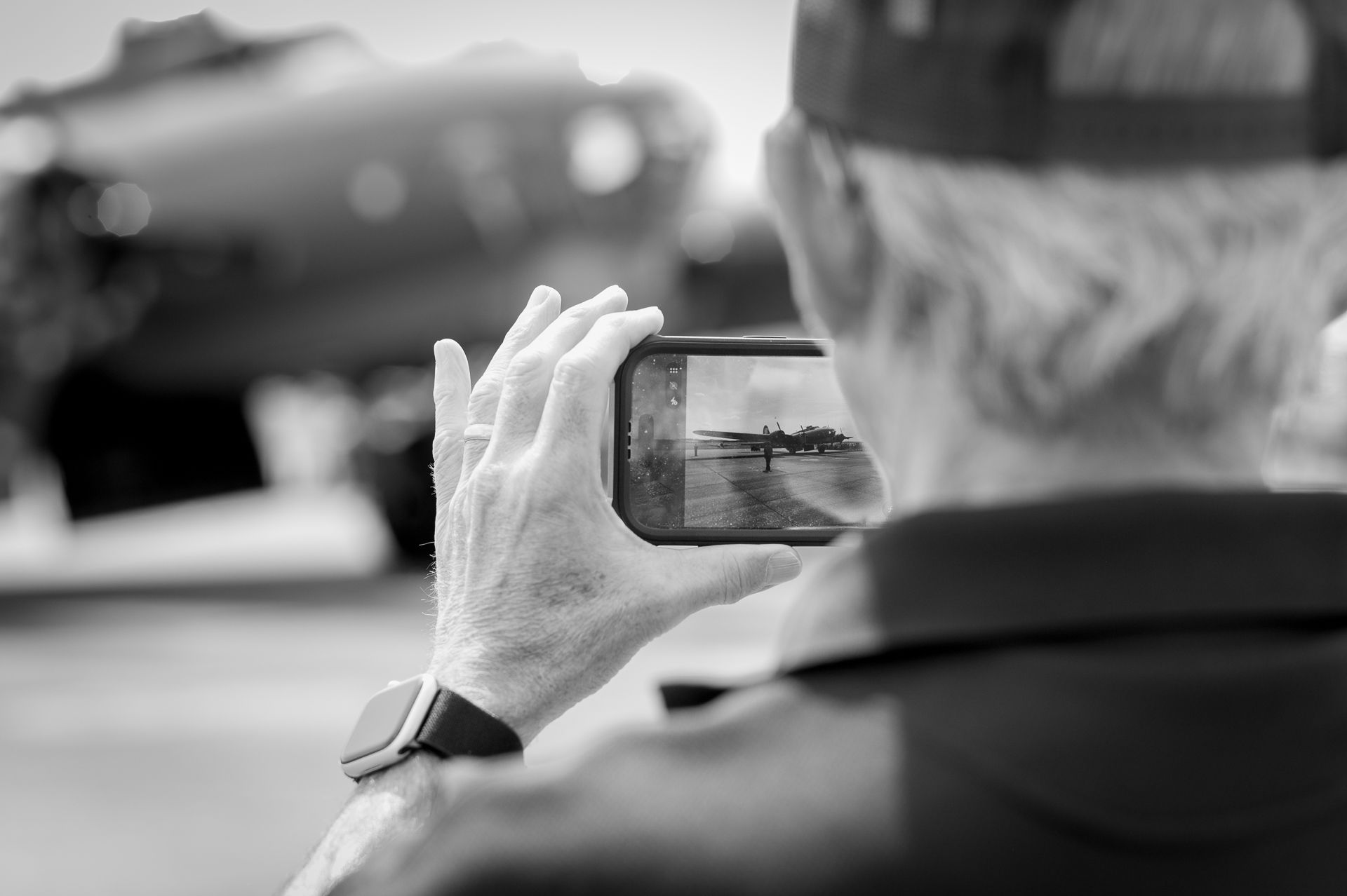Elderly man photographing a vintage WWII aircraft with smartphone, captured in black and white at an airshow.