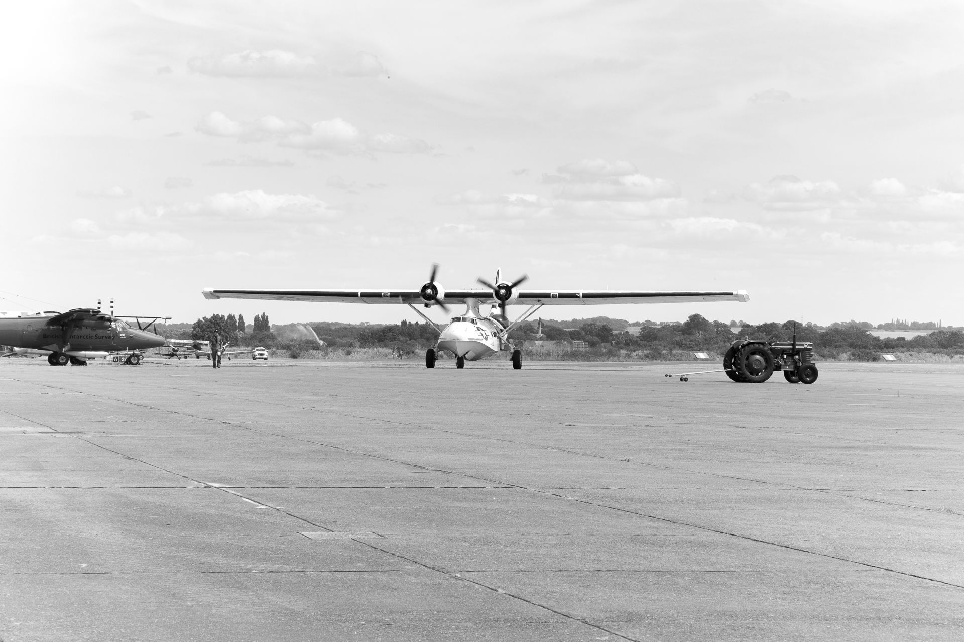 Vintage black and white photo of a twin-engine propeller aircraft taxiing on an airfield tarmac with a tractor nearby.