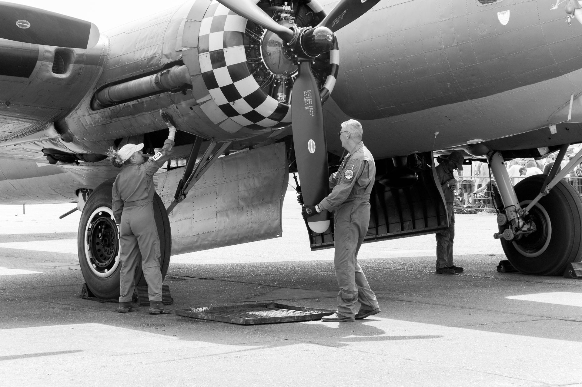 WWII-era B-17 bomber on tarmac with ground crew inspecting checkered nose engine in black and white photo.
