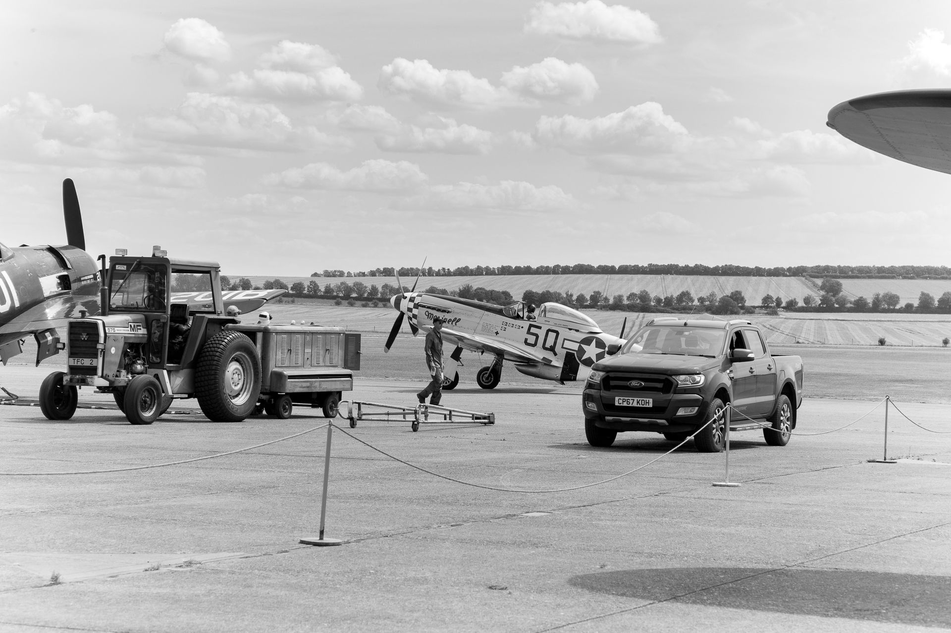 Vintage WWII aircraft on tarmac with tractor and pickup truck at airshow, black and white photography.