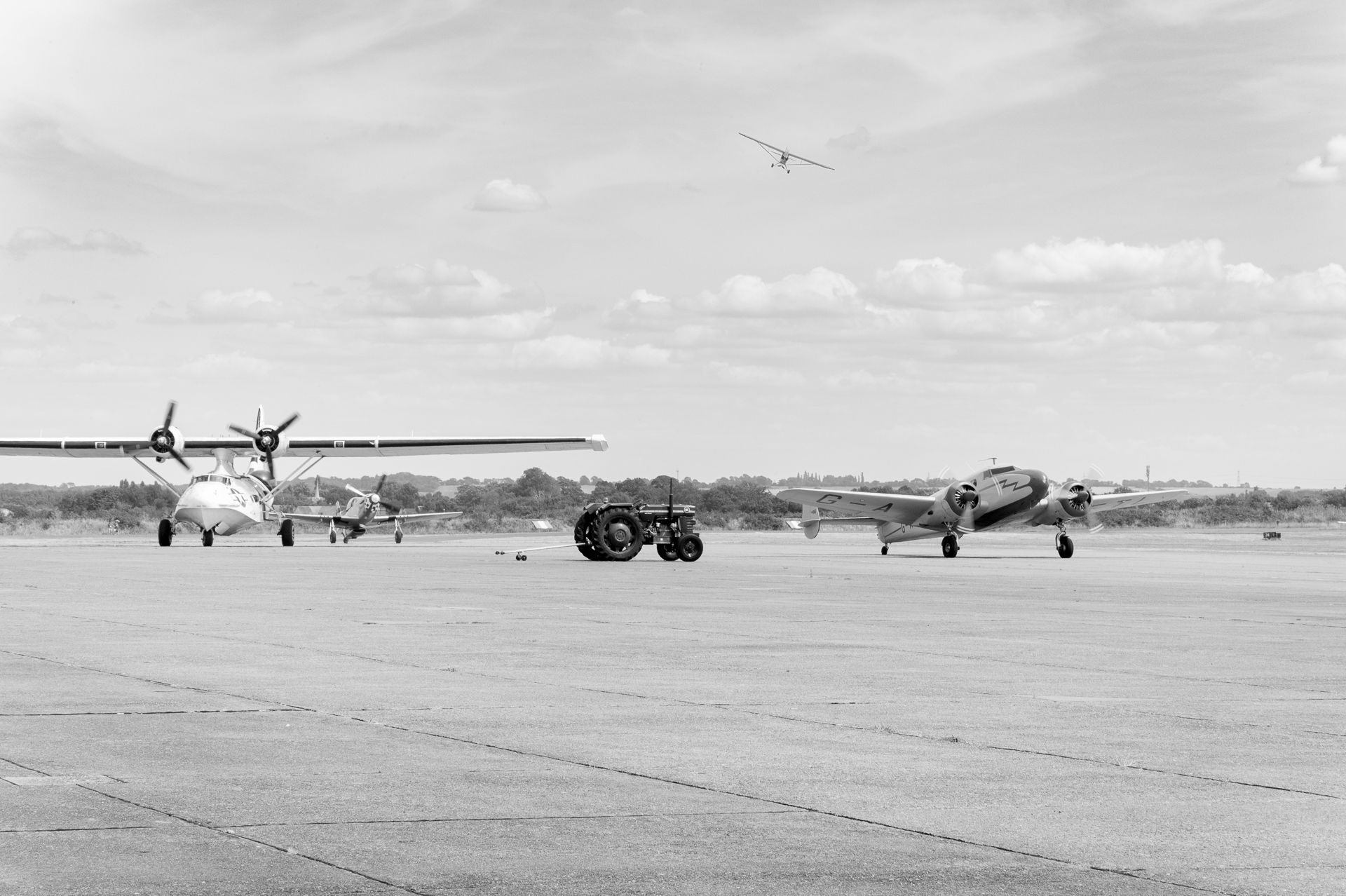 Vintage black and white photo of propeller aircraft and ground vehicles on an airfield tarmac under a cloudy sky.