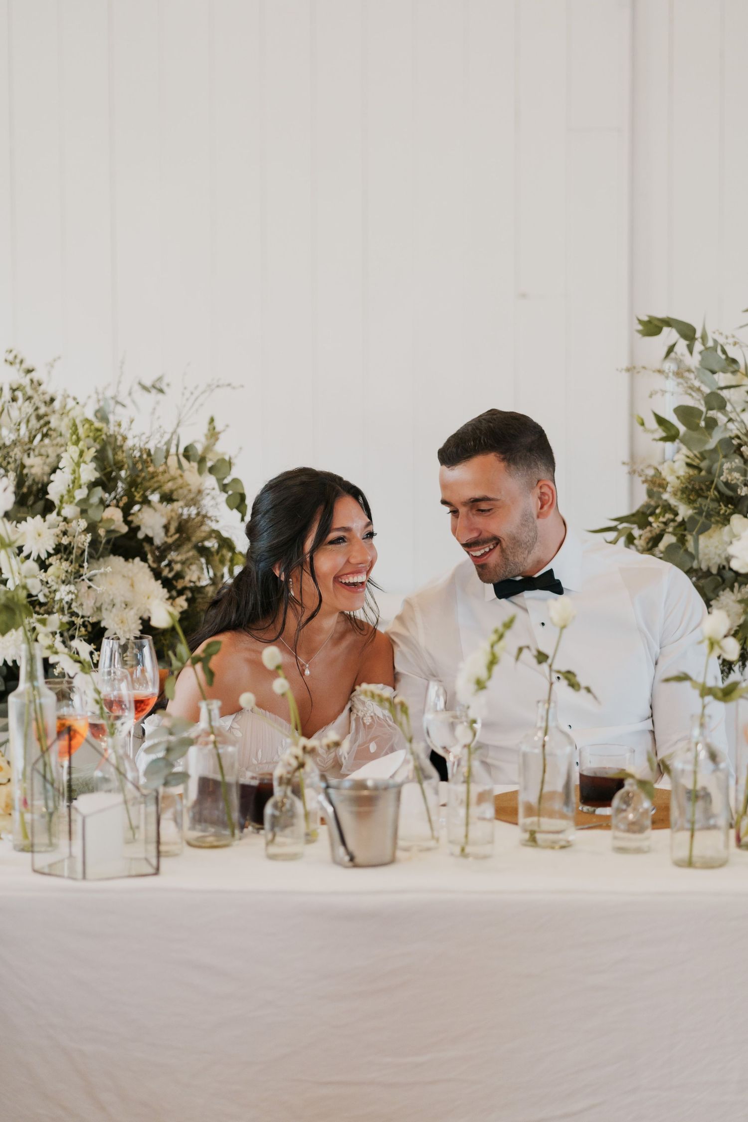 Bride and groom share a joyful laugh at their elegantly decorated sweetheart table with white floral centerpieces.