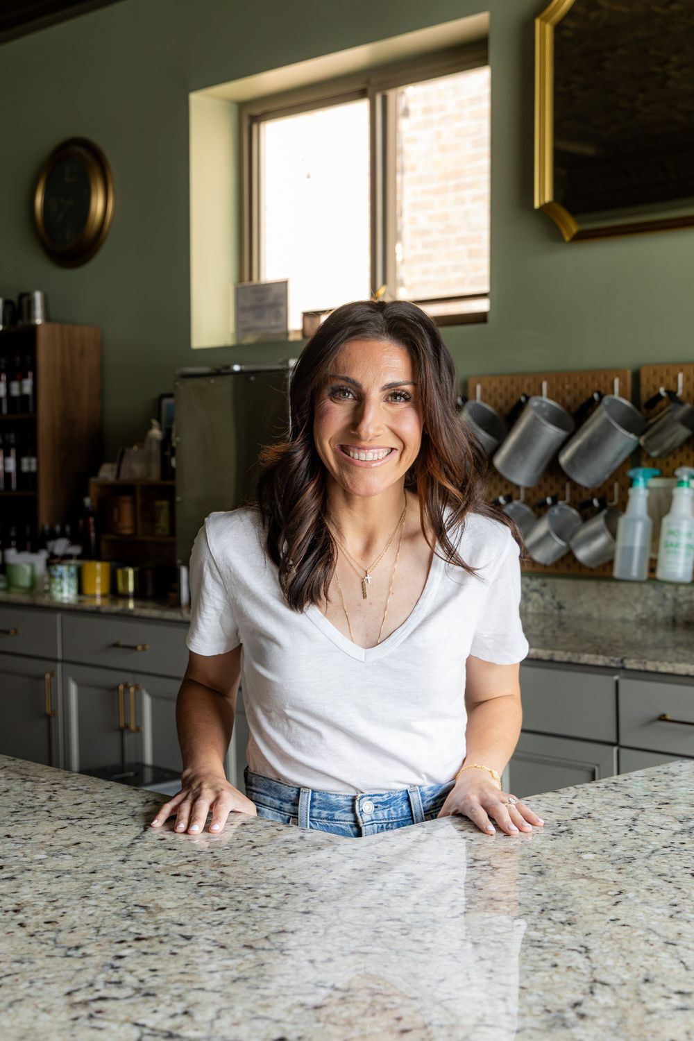 Smiling brunette woman in white t-shirt and jeans leaning on granite kitchen counter in modern green kitchen.