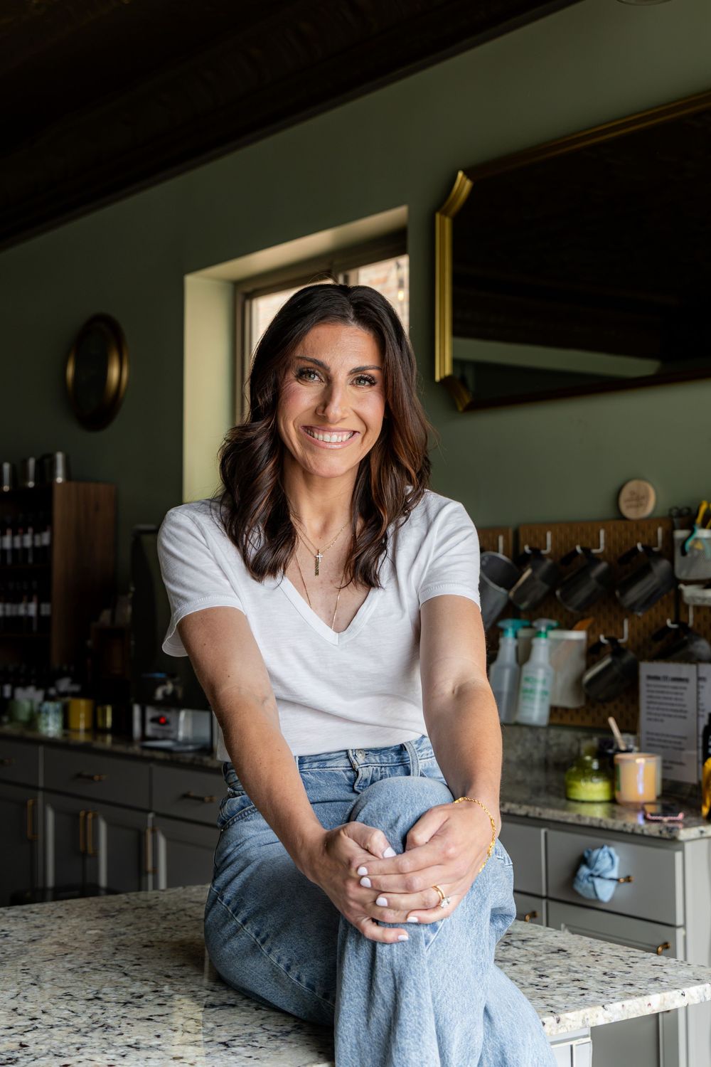Smiling woman in white t-shirt and jeans sitting on kitchen counter with green cabinets in background.