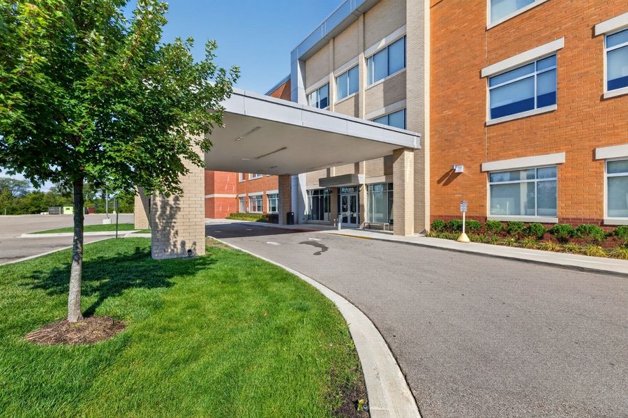 Modern brick building entrance with covered driveway, manicured lawn, and trees under a clear blue sky.