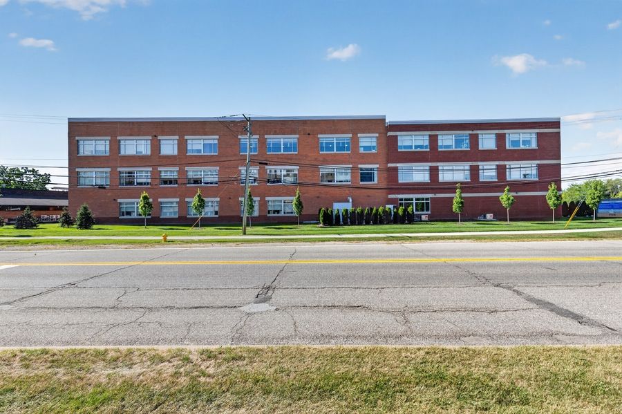 Three-story red brick apartment building along a wide road with green lawn and trees on a sunny day.