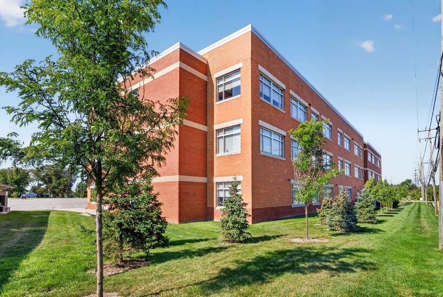 Three-story red brick office building with large windows surrounded by young trees and green lawn on a sunny day.