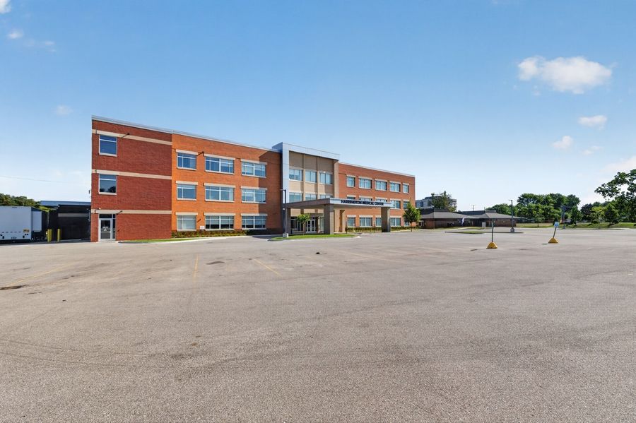 Three-story brick school building with large parking lot under blue sky, featuring modern entrance and adjacent single-story wing.