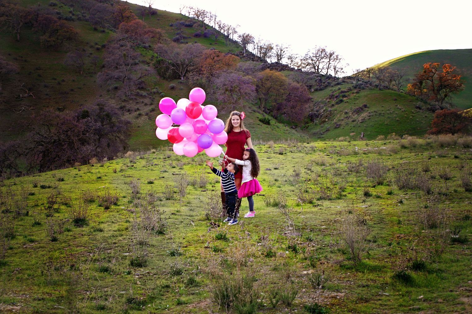 A woman and two children hold a bunch of pink balloons on a green grassy hillside with autumn trees in the distance.