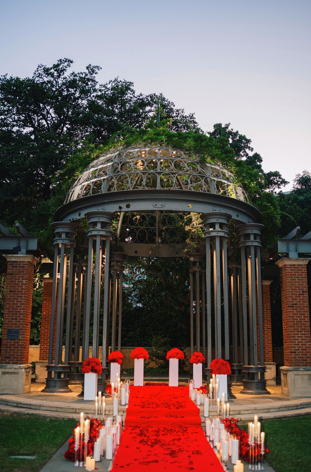 Elegant gazebo wedding ceremony setup with red carpet aisle, white pillar candles, and red floral arrangements at dusk.