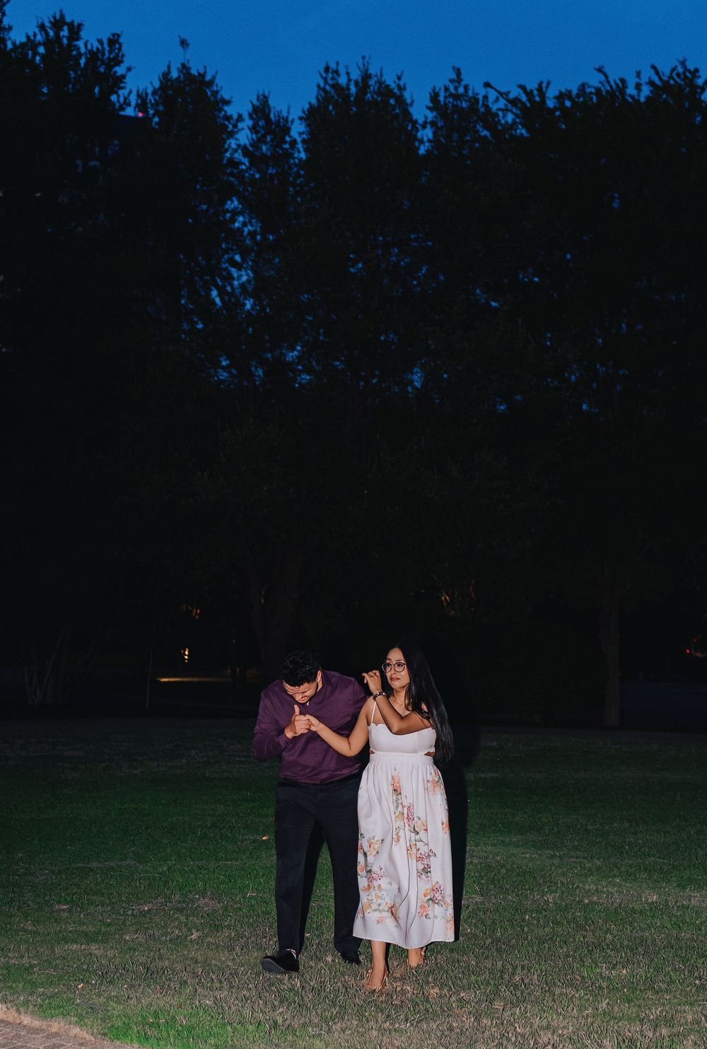 Couple sharing a romantic moment at dusk in a park, man in purple shirt kisses woman's hand in white floral dress.
