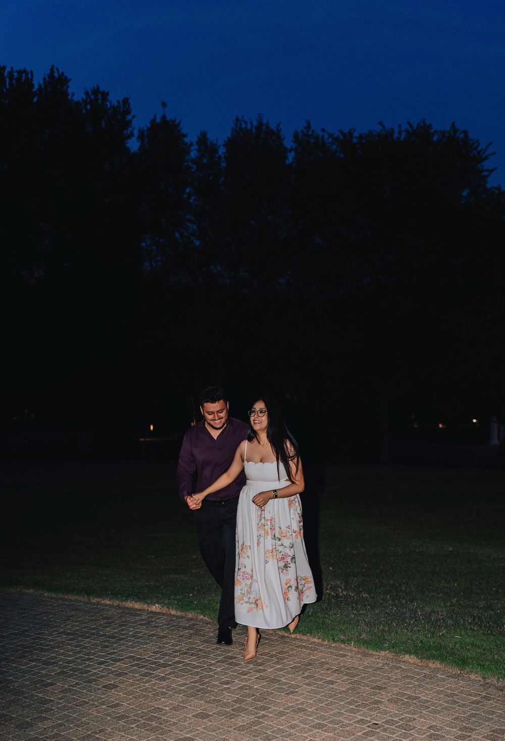 Couple walking hand in hand on a brick path at dusk, surrounded by tall trees under a deep blue evening sky.