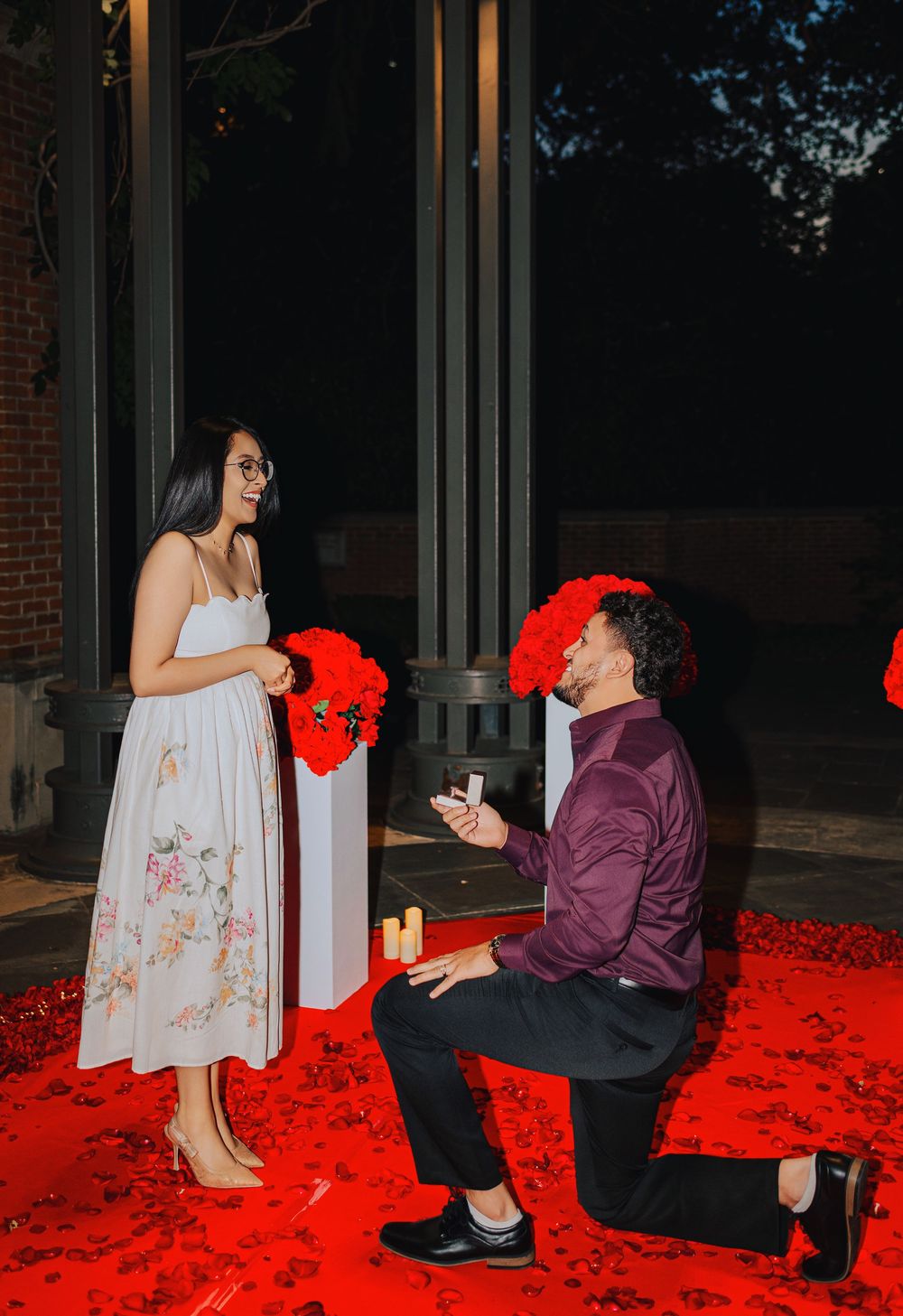 Man kneeling on red rose petal carpet proposing with ring box to woman in white floral dress at romantic night setting.