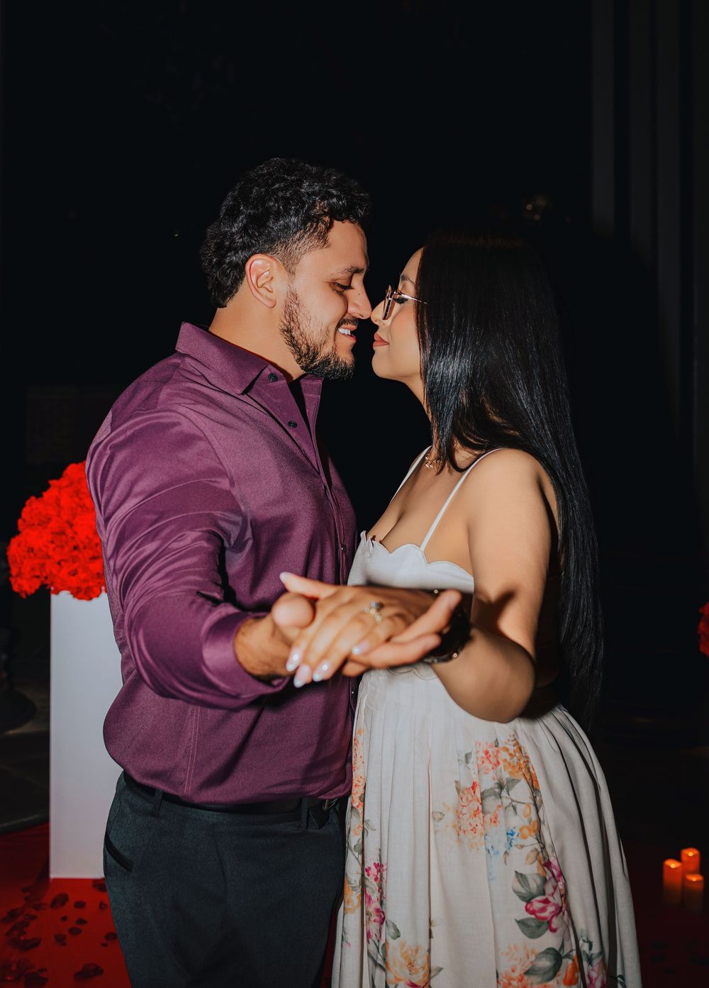 Couple sharing a romantic kiss during an engagement proposal surrounded by red roses and candles.