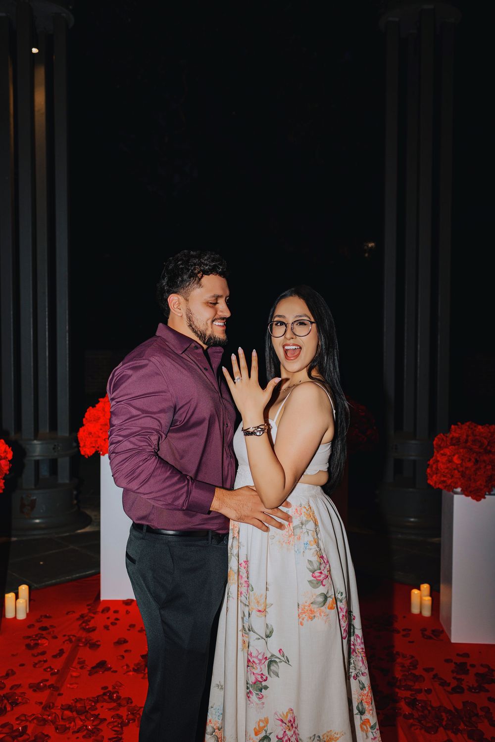 Couple celebrating engagement at night with red roses, candles, and columns, woman showing ring while smiling.