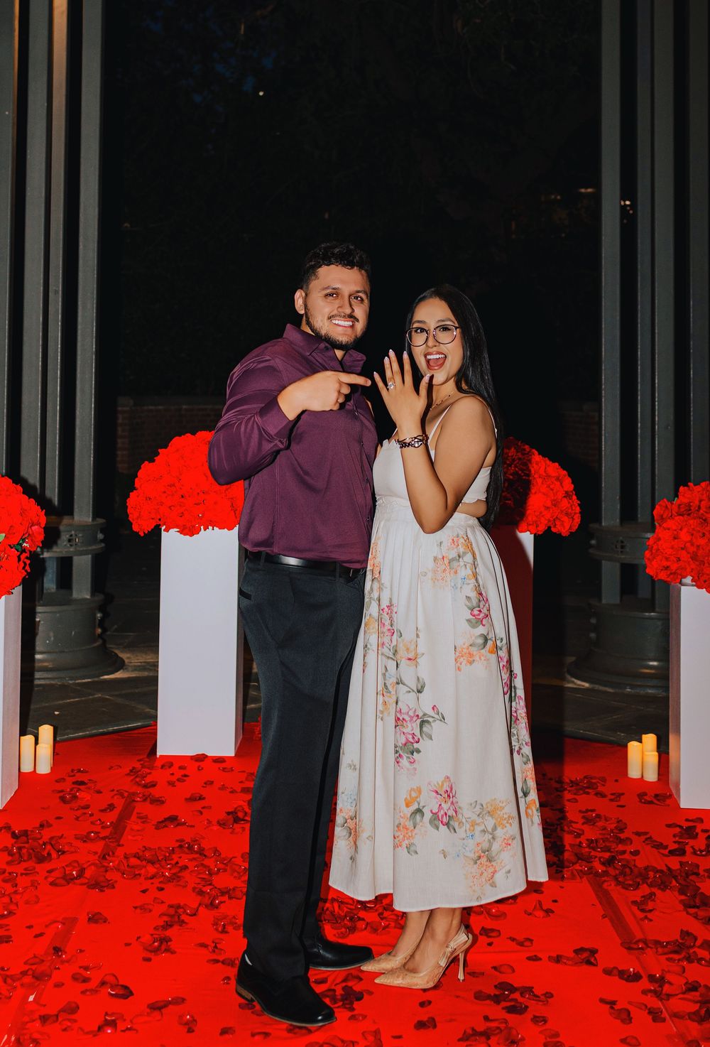 Happy couple celebrating engagement showing off ring on red petal carpet with floral decorations.