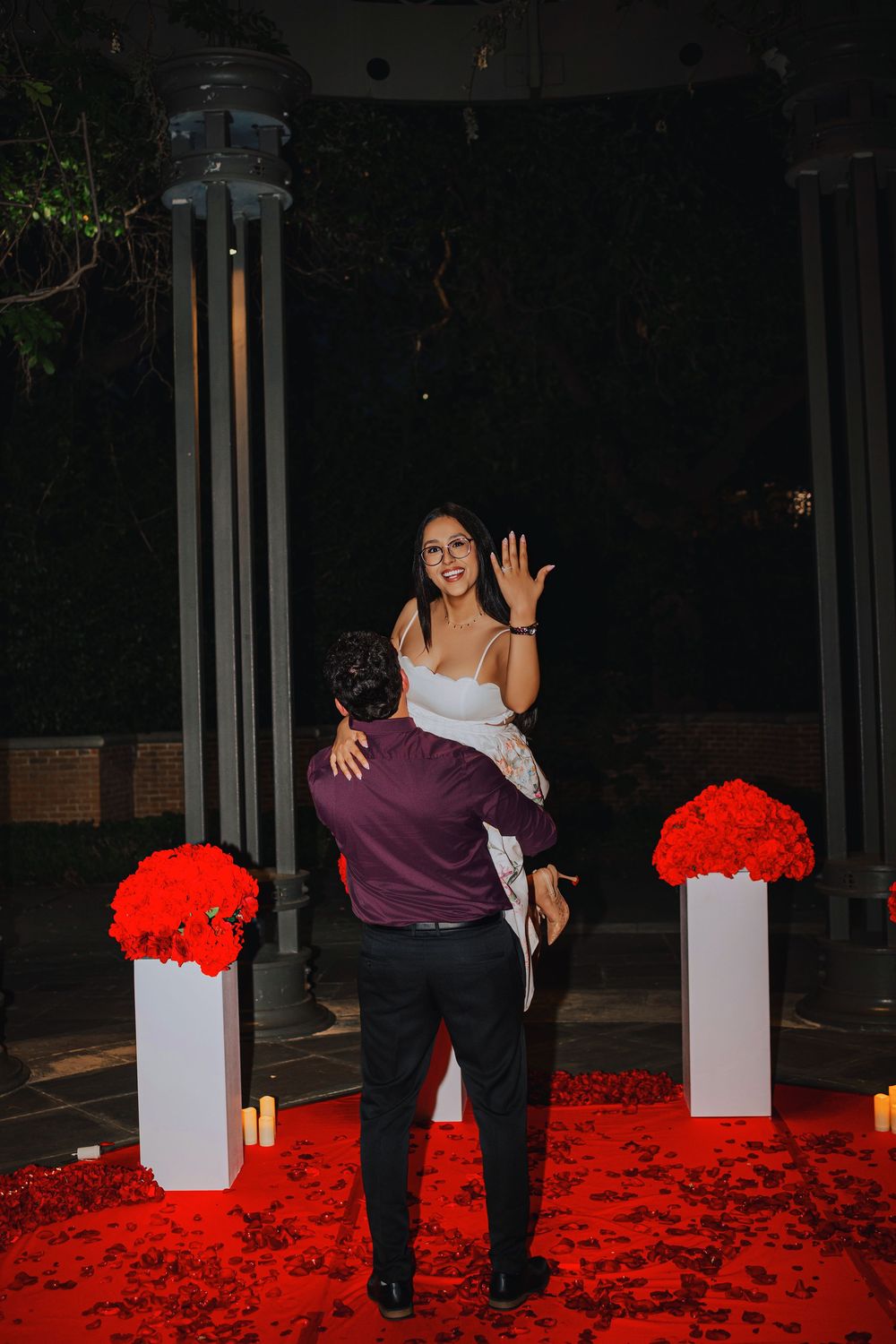 Man lifts woman in white dress during romantic red rose petal proposal with candles and floral arrangements at night.