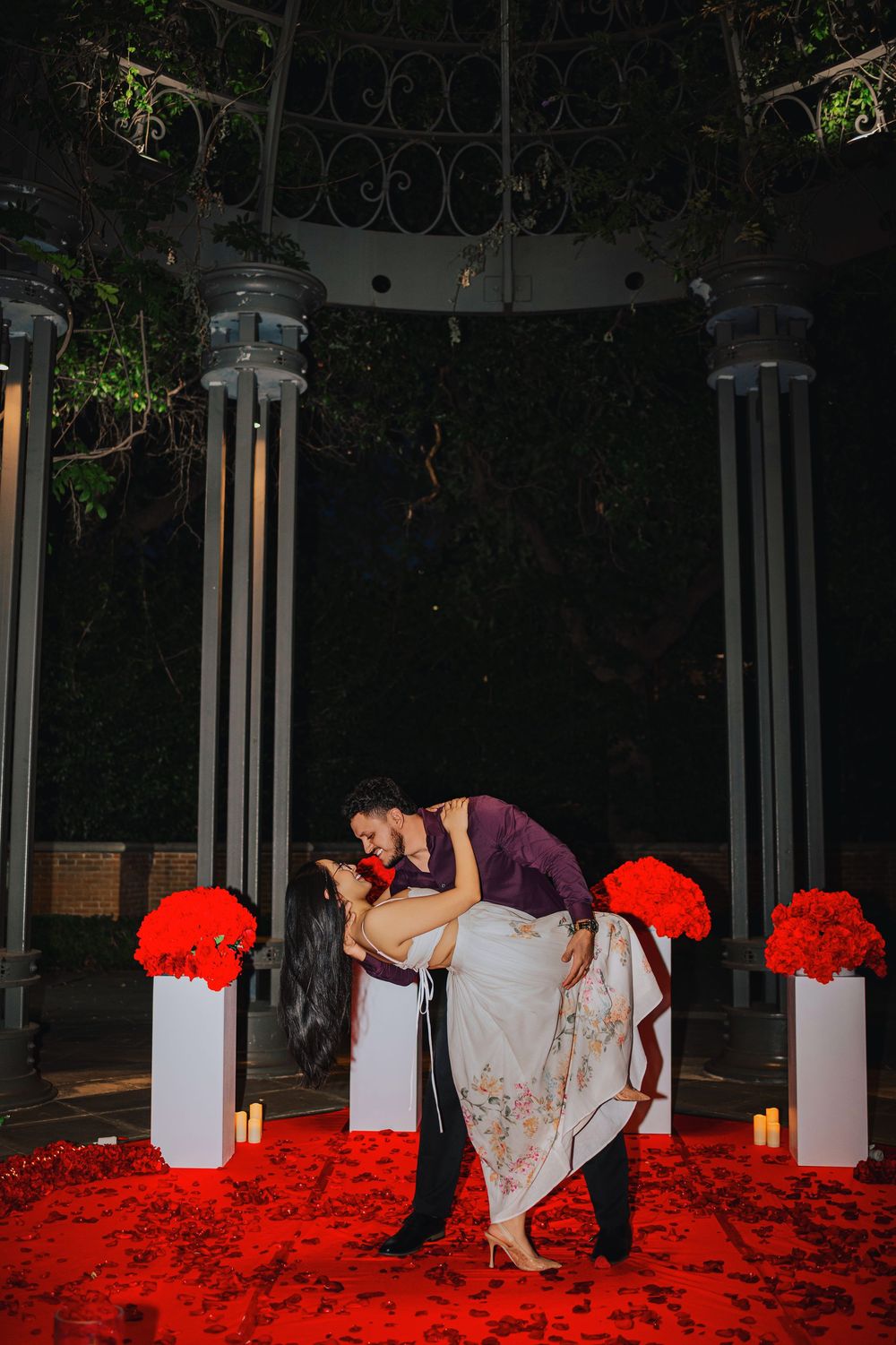 Couple sharing a romantic dip kiss under a gazebo surrounded by red rose petals and floral arrangements at night.