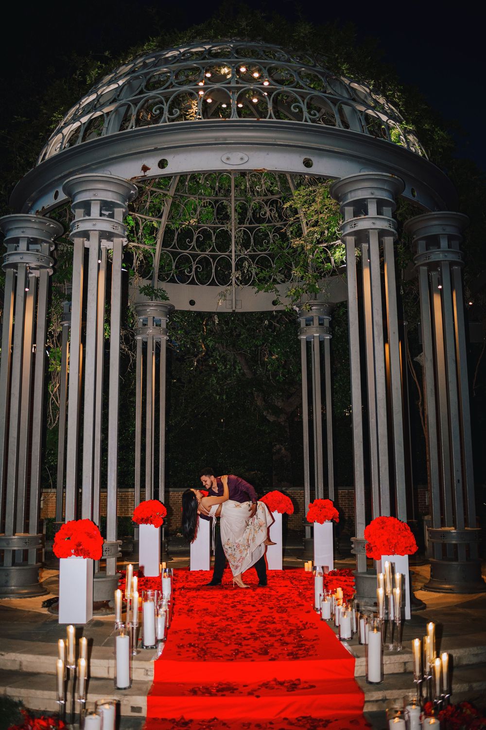 Couple shares a dip kiss under elegant gazebo on red rose petal aisle with candles and red floral arrangements at night.