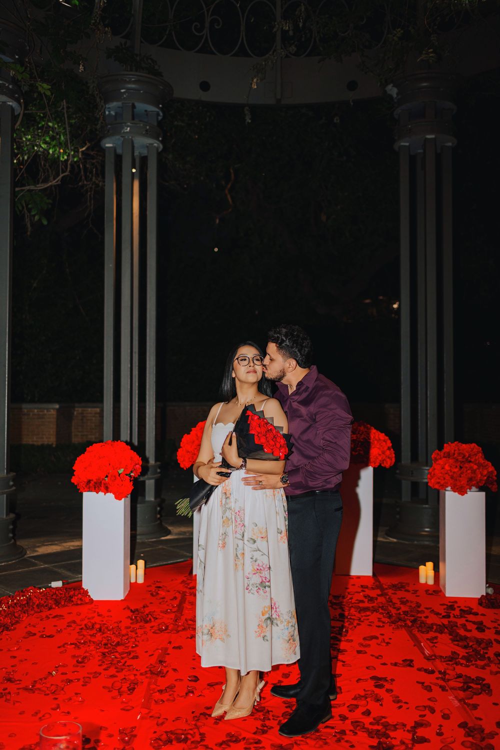 Romantic couple embracing under a gazebo at night, surrounded by red rose petals and floral arrangements.