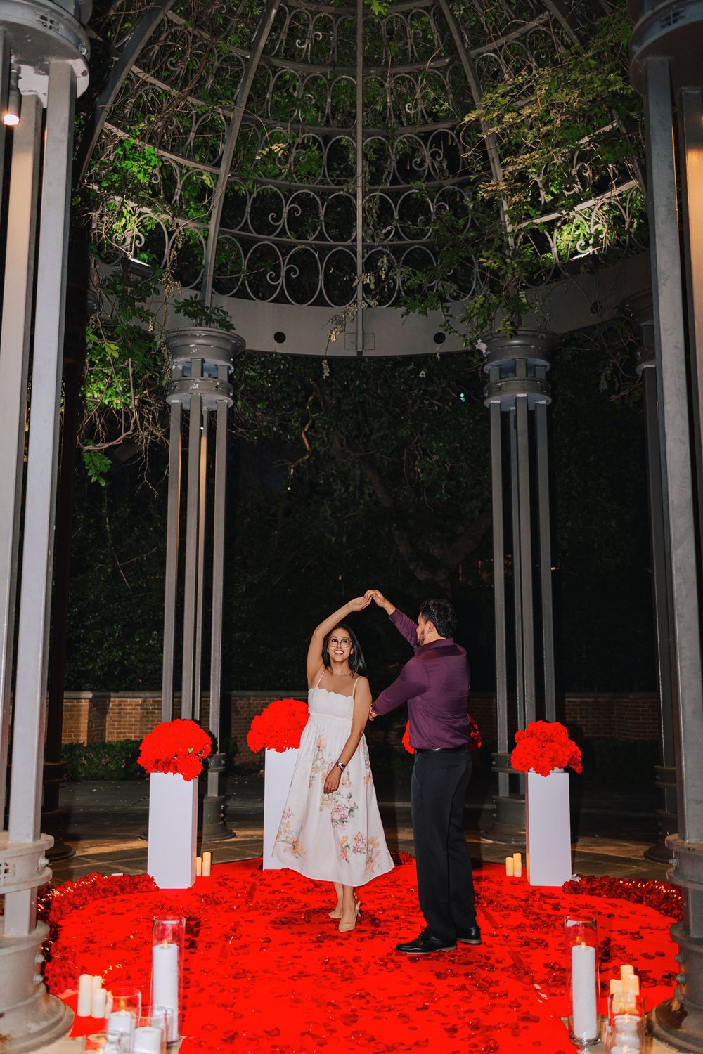 Couple dancing under an ornate gazebo surrounded by red rose petals, candles, and floral arrangements at a romantic night proposal.