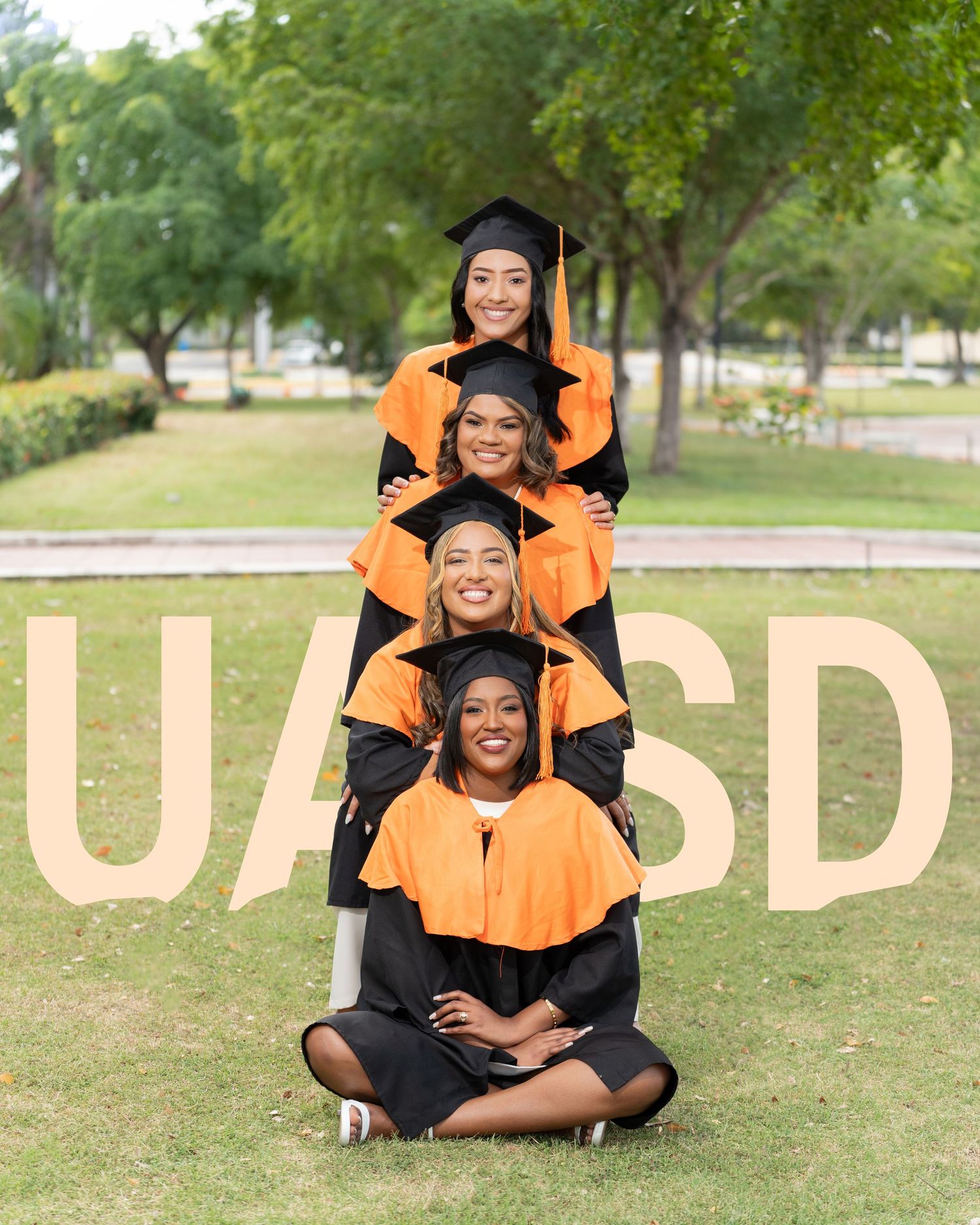 Four UASD graduates in orange and black caps and gowns pose together outdoors on green grass.
