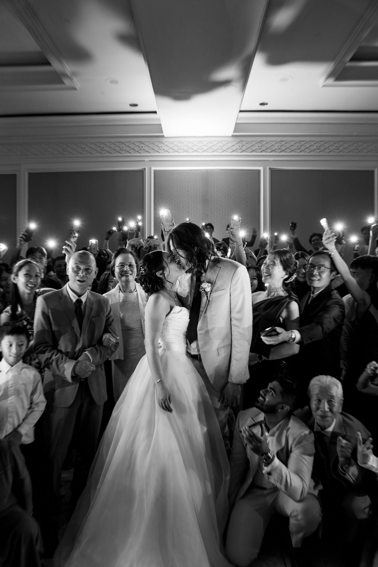 Black and white photo of a bride and groom kissing surrounded by guests holding up phone lights at their wedding reception.