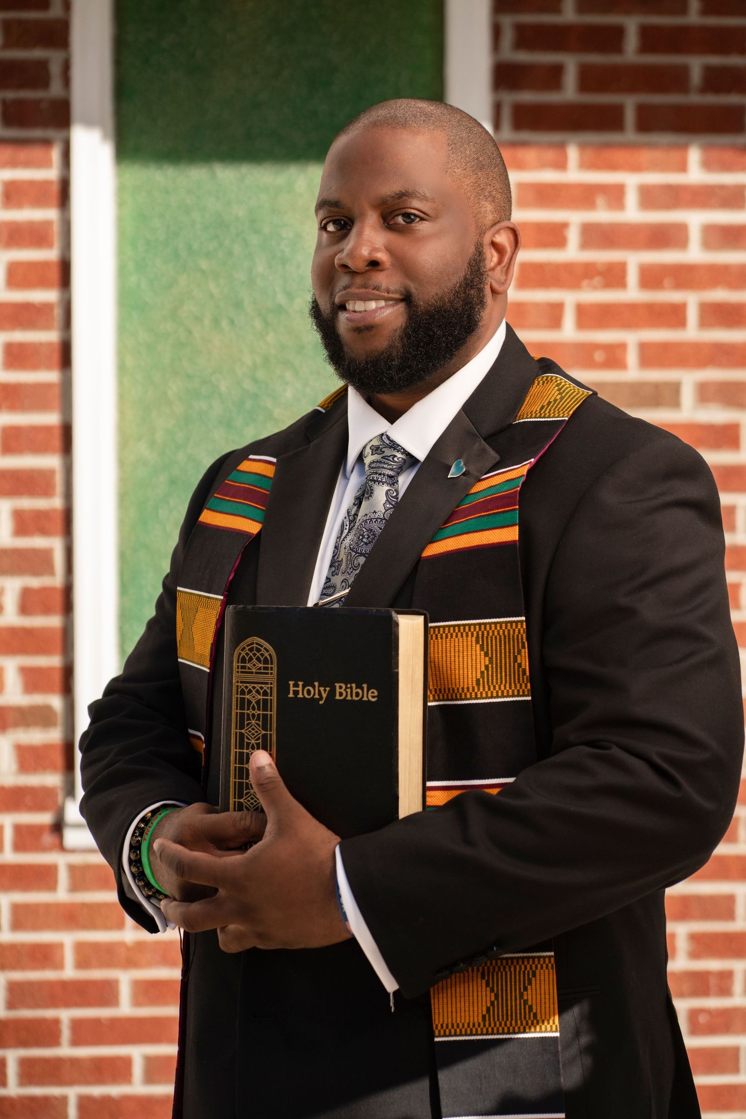 A bearded Black man in a suit with a kente cloth stole holds drumsticks, posing confidently for a portrait.