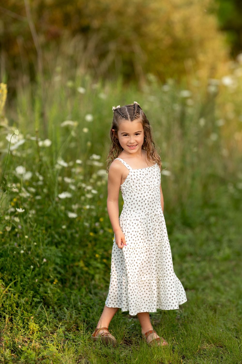 Smiling young girl in a white polka dot sundress standing in a green meadow with wildflowers.