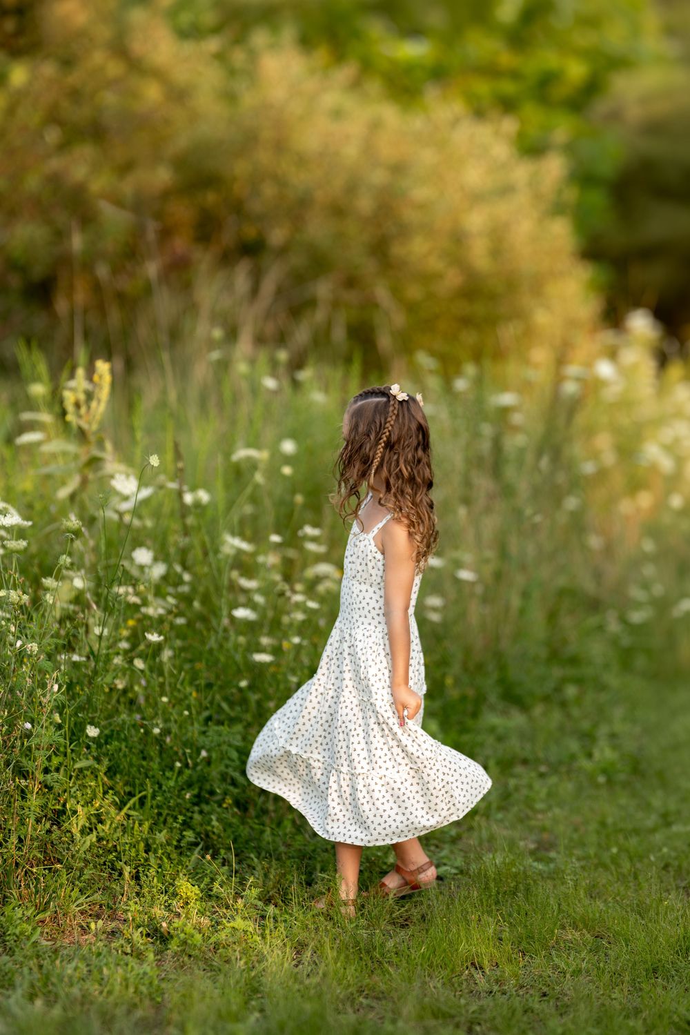Young girl in white polka dot dress twirling in a lush green meadow with wildflowers on a sunny day.