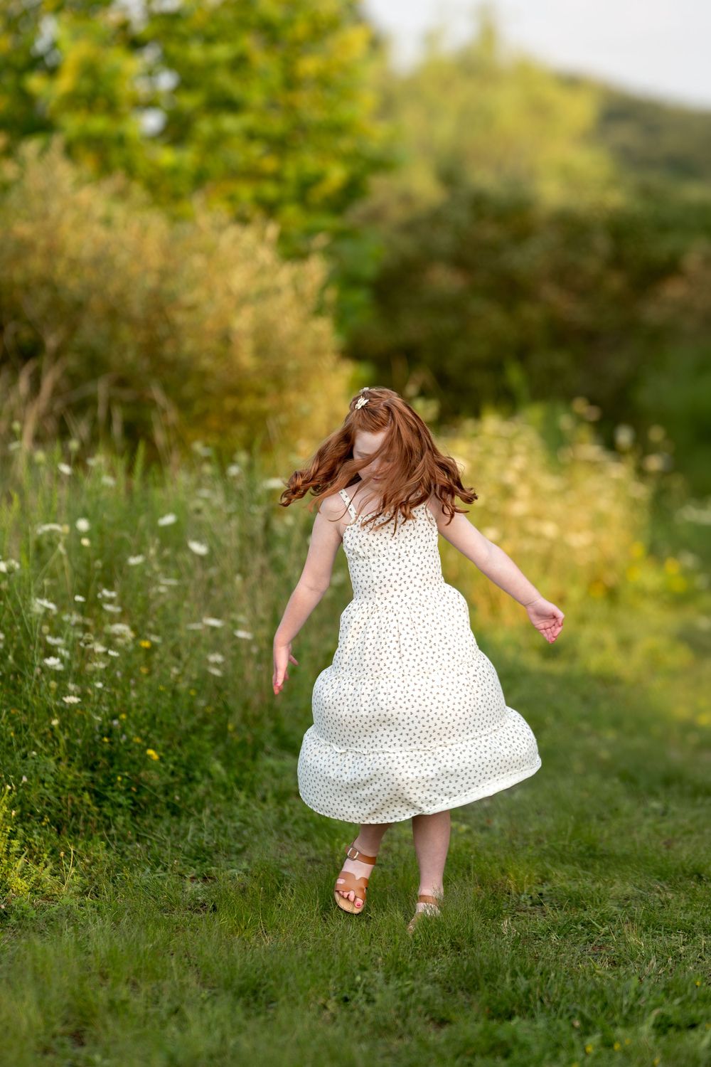 Redhead girl in white dress twirling joyfully in a sunny green meadow.