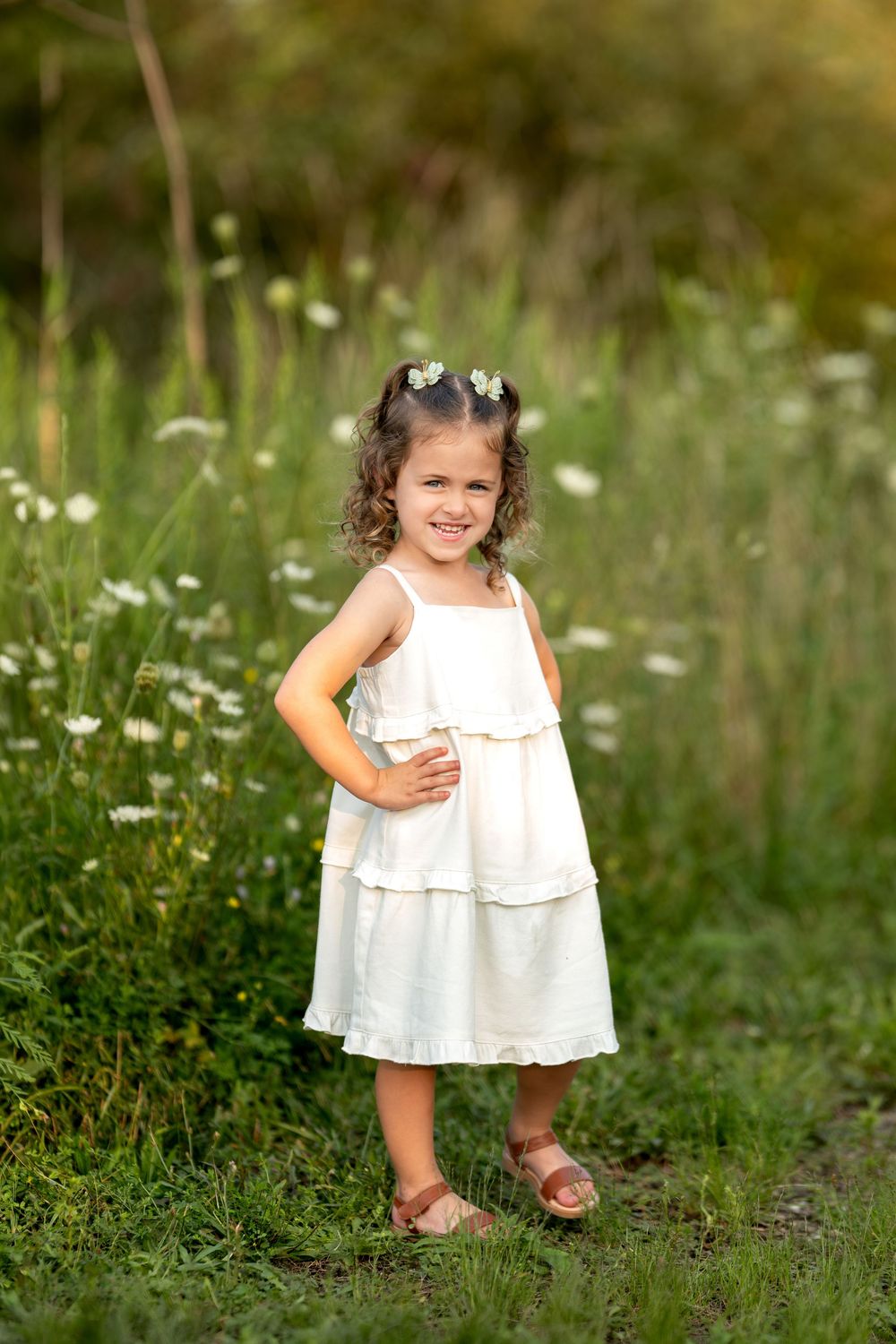 Happy young girl in white ruffled sundress posing with hand on hip in a green meadow.