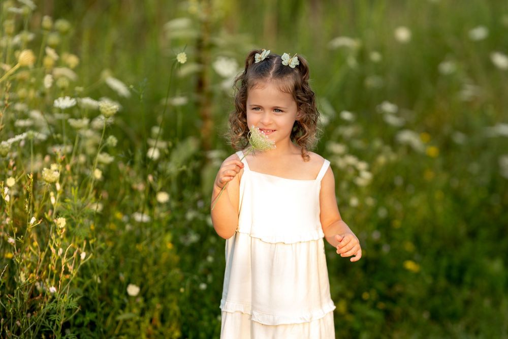 Young girl with flower crown in white dress holding a daisy in a meadow.