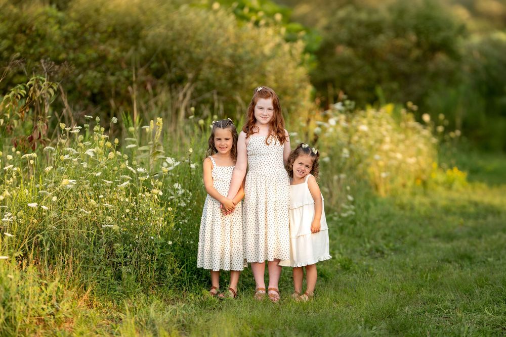 Three sisters in white dresses posing together in a sunny wildflower meadow.