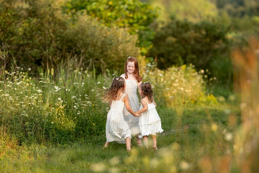 Three girls in white dresses holding hands and playing in a wildflower field.