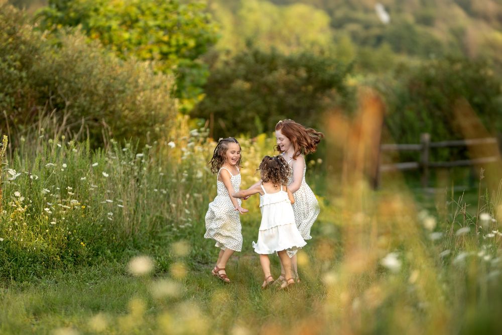 Three girls in white dresses holding hands and dancing in sunny meadow.