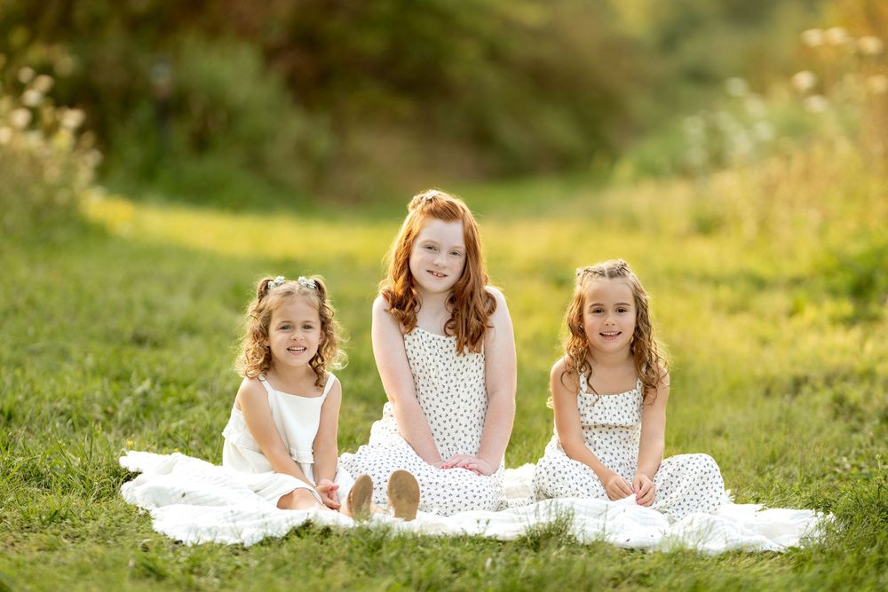 Three sisters in white dresses sitting on a blanket in a sunny green field.
