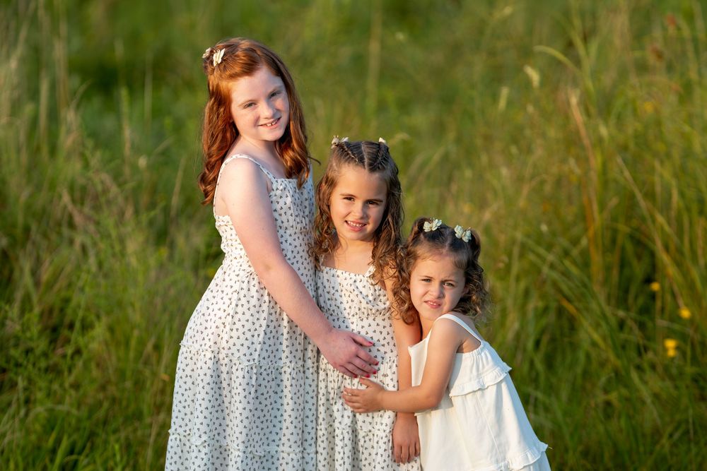 Three girls in white dresses pose together in a sunny green field, smiling warmly in natural golden light.