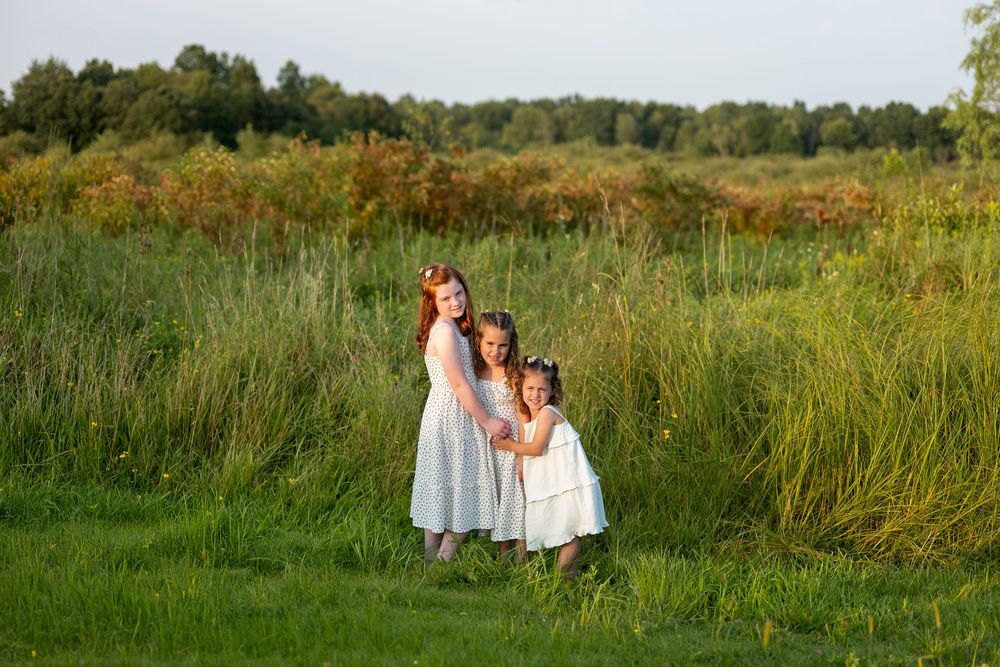 Three young girls in white dresses pose together in a sunny green meadow.