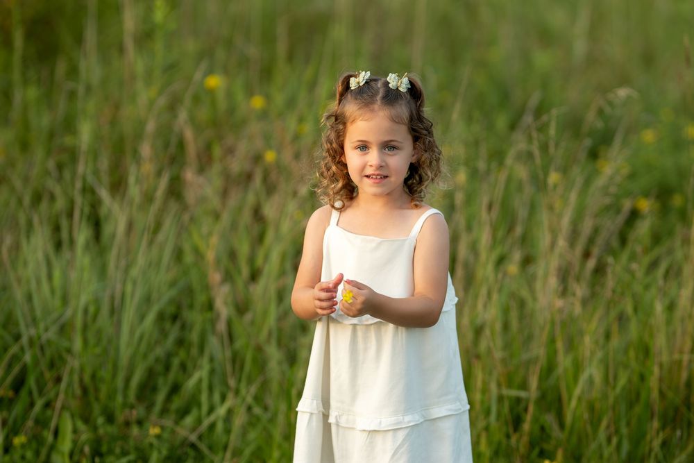 A young girl in a white dress with flower hair accessories smiles while standing in a lush green meadow.