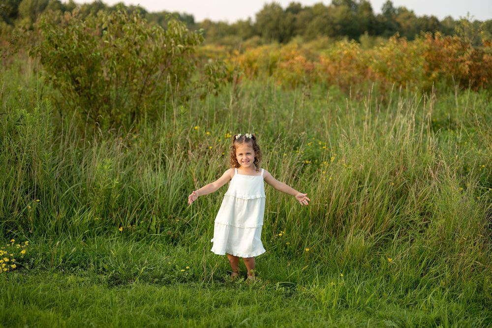 A young girl in a white dress runs through a lush green meadow with arms outstretched on a sunny day.