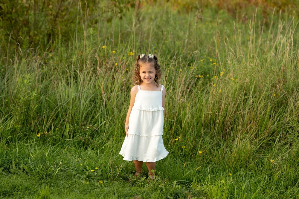 A young girl in a white dress stands smiling in a lush green meadow with wildflowers on a sunny day.