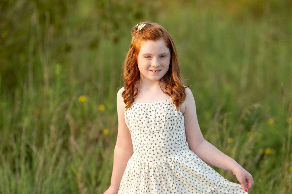 A smiling redhead girl in a white polka dot dress poses in a sunny green meadow with wildflowers.