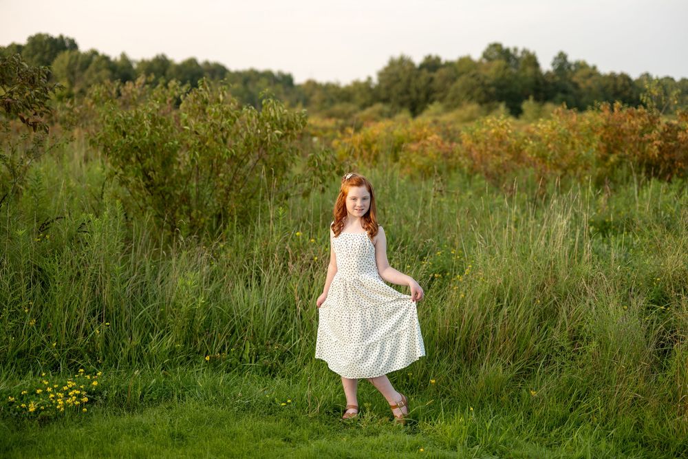 Red-haired girl in white dress twirling in lush green field at sunset.