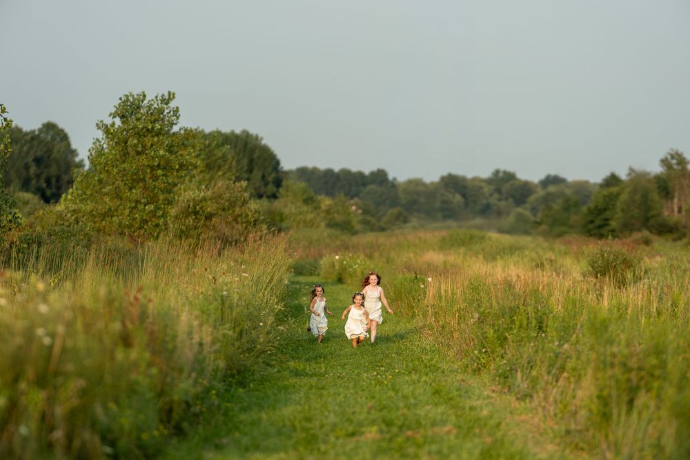 Two girls in white dresses running through a lush green meadow path on a sunny day.